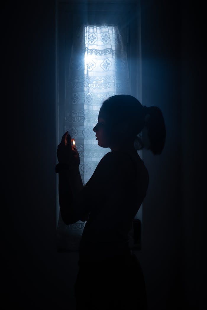 Artistic silhouette of a woman holding a candle, illuminating a dark room by the window.